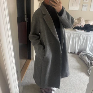 Grå lång oversized kappa - Säljer en stilren grå kappa i längre modell med klassisk krage och knäppning framtill. Jackan har en rak passform och är tillverkad i ett ull-liknande material. Perfekt för dig som gillar en clean och tidlös look.