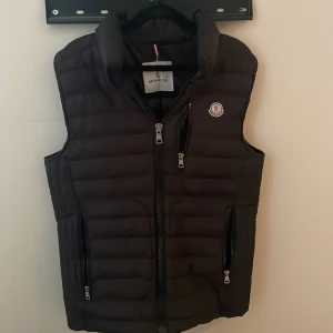 Svart dunväst från Moncler - Svart dunväst från Moncler med quiltad design och hög krage. Västen har två sidofickor med dragkedja, en bröstficka med dragkedja och Moncler-logga på bröstet. Snygga silverfärgade detaljer på dragkedjorna och insida med serietidningsmönster.