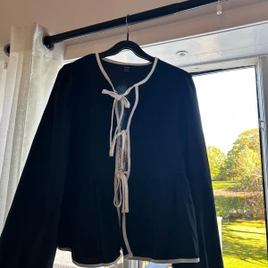 Svart kofta med vita knytband - Svart kofta/blus från shein med vita kanter och tre dekorativa knytband framtill. Långärmad och lätt material, perfekt för lager-på-lager. Snygg kontrast mellan svart och vitt, ger en stilren och trendig look.