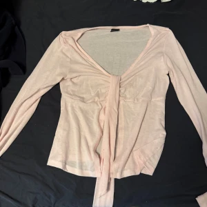 Ljusrosa topp med knyt - Söt ljusrosa topp med långa ärmar och knyt framtill. Tunn och lätt i materialet, perfekt för lager-på-lager. Djup v-ringning ger en feminin touch och toppen har en smickrande passform.