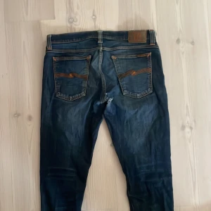 Mörkblå jeans från Nudie Jeans - Säljer ett par mörkblå jeans från Nudie Jeans i storlek 32/34. Skicket är bra förutom hål i ett av knäna, i skrevet och i en av fickorna. Hålet i knäet är mest snyggt enligt mig.
