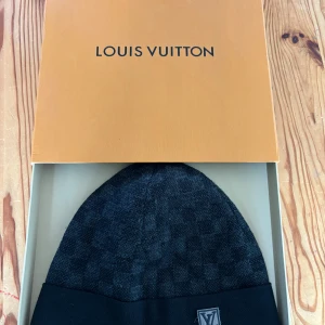 Louis Vuitton mössa  - Exklusiv svart mössa från Louis Vuitton med diskret rutigt mönster och uppvikt kant. LV-logga i grått framtill ger en lyxig touch. Stickad i mjukt material som känns skönt mot huden. Perfekt för dig som vill ha en stilren och ikonisk accessoar.