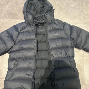 Svart dunjacka Peak Performance XL - Säljer en svart dunjacka från Peak Performance i storlek XL. Jackan har en puffig design, huva och dragkedja framtill. Perfekt för kalla dagar och har en stilren look med diskret logga på bröstet. Klassisk modell som håller värmen riktigt bra.