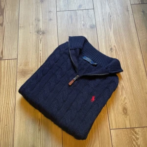 Mörkblå stickad tröja Polo Ralph Lauren - Mörkblå kabelstickad tröja från Polo Ralph Lauren med halv dragkedja och röd broderad logga på bröstet. Tröjan har hög krage, ribbade muddar och är långärmad. Perfekt för dig som gillar klassisk och stilren look.
