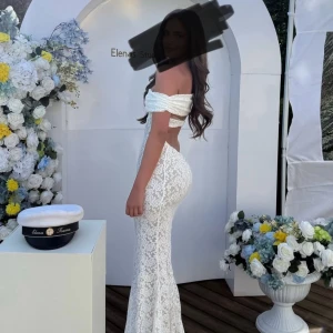 Vit spetsklänning offshoulder - Supersnygg lång vit spetsklänning med offshoulder och figurnära passform. Klänningen har öppen rygg och vackra spetsdetaljer över hela tyget. Perfekt för student eller sommarens speciella tillfällen.