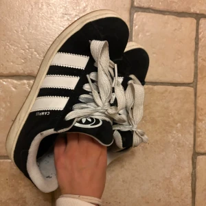 Adidas Campus sneakers svart/vit - Svarta Adidas Campus sneakers med klassiska vita ränder och vit sula. Skorna har snörning och är gjorda i mocka med detaljer i syntet. Perfekta för dig som gillar en clean och tidlös streetstil.