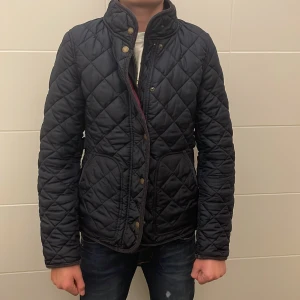 Svart quiltad jacka från GANT - Quiltad svart jacka från GANT med ståkrage och snygga guldfärgade knappar. Jackan har dragkedja och tryckknappar framtill, två fickor och en mörkröd insida. Nypris ~ 1800. Mitt pris - 319. Originell storlek är S men den sitter som xs.