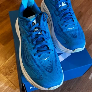 Blå Hoka Clifton 10 sneakers - Provat bara på gymmet 1 gång -  Säljer ett par Hoka Clifton 10 sneakers i en fräsch blå färg med vit sula. Skorna har ventilerande mesh-ovandel, snörning och en riktigt skön platt sula som passar perfekt för löpning eller träning. Märket syns tydligt på sidan och tungan. Nypris 2200:-    mitt pris : 1250:- 