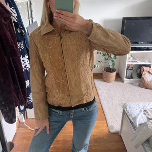 Beige mockajacka med pärldetaljer S - Snygg beige mockajacka med dragkedja och coola pärldetaljer framtill. Jackan har klassisk krage, lång ärm och är tillverkad i äkta skinn med polyesterfoder. Perfekt för dig som vill sticka ut med en unik och trendig look.
