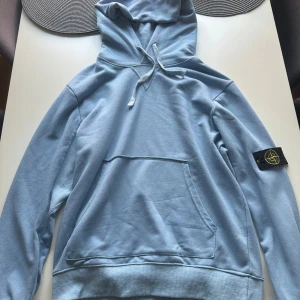 Ljusblå hoodie från Stone Island - Snygg ljusblå hoodie från Stone Island med klassisk patch på ärmen. Tröjan har huva med snörning, stor magficka och ribbade muddar. Perfekt för dig som gillar streetwear och vill ha något bekvämt och stilrent. Materialet är mjuk bomull.