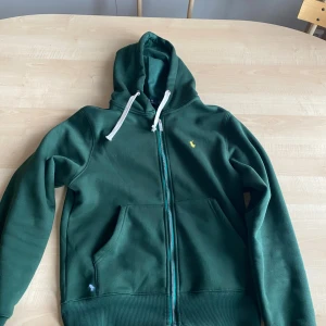 Mörkgrön hoodie från Polo Ralph Lauren - Mörkgrön hoodie från Polo Ralph Lauren med gul broderad logga på bröstet. Tröjan har huva med vita snören, två fickor fram och hel dragkedja med Polo-detalj. Mjuk insida i fleece och ribbade muddar. Perfekt för chill dagar och streetstyle.