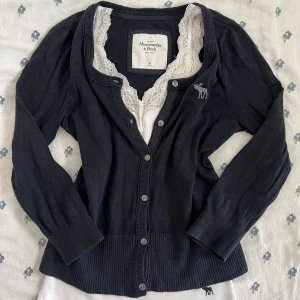 Mörkblå kofta från Abercrombie & Fitch - Mörkblå kofta från Abercrombie & Fitch med rund halsringning och knäppning framtill. Broderad älg-logga på bröstet och bred ribbad kant nedtill och vid ärmslut. Långärmad och i mjukt material, perfekt för lager-på-lager. ¾ längd armar. * linnetoppen ingår ej och är inte till salu * Ungefärliga mått (platta): Axel till axel: 33 cm Axel till botten: 50 cm Armhåla till armhåla: 39 cm Axel till handled: 50 cm