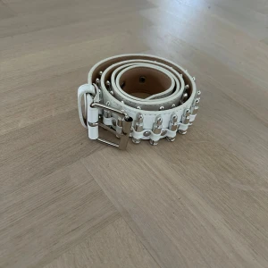 Vitt bälte med silvriga nitar - Snyggt vitt bälte i skinn med silvriga nitar längs hela framsidan. Bältet har en klassisk metallspänne och flera hål för justerbar passform. Insidan är beige och bältet har en cool, edgy vibe som lyfter vilken outfit som helst.