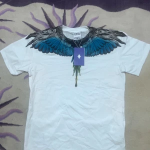 Vit t-shirt med blå vingar Marcelo Burlon - Snygg vit t-shirt från Marcelo Burlon County of Milan med ikoniskt tryck av blå och svarta vingar runt halsen. Klassisk passform och rund hals. Perfekt statement-plagg för dig som vill sticka ut. Materialet är mjuk bomull.