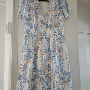 Blommig sommarklänning med volang - Superfin vit klänning med blått och orange blommönster, v-ringning och volangdetaljer. 