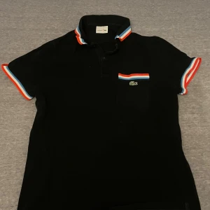 Svart Lacoste piké med färgglada detaljer - Svart pikétröja från Lacoste med korta ärmar och krage. Snygga ränder i orange, vit och blå på ärmslut, krage och bröstficka. Klassisk Lacoste-logga på fickan. Tröjan är gjord i mjuk bomull och har en normal passform.