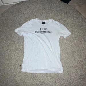 Vit Peak Performance t-shirt - Vit t-shirt från Peak Performance med svart tryck på bröstet. Klassisk rund halsringning och korta ärmar. Tillverkad i mjuk bomull för en skön känsla. Perfekt basic-plagg med stilren design och logga framtill.