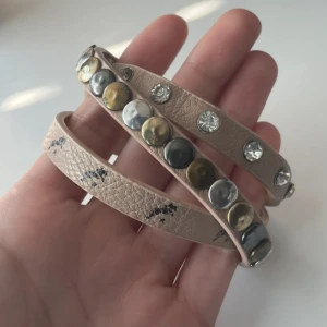 Beige läderarmband med nitar och stenar - Coolt beige armband i läderimitation som snurras flera varv runt handleden. Prydd med blanka nitar i silver och guld samt glittrande stenar. Perfekt accessoar för att ge din outfit en edgy touch.