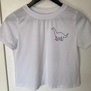 Vit t-shirt med dinosaurietryck - Vit t-shirt i bomull med korta ärmar och en enkel dinosaurie-illustration tryckt på bröstet. Modellen är rak och har en klassisk rund halsringning. Perfekt för dig som gillar minimalistiska och lekfulla detaljer.