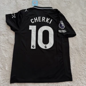 Manchester City svart fotbollströja Puma - Snygg svart Manchester City fotbollströja med CHERKI och nummer 10 på ryggen. Tröjan har vita detaljer, korta ärmar och klassisk krage. Premier League-logga och 'No Room for Racism' på ärmen, samt Etihad Airways tryck på bröstet. Tillverkad i lätt polyester med Drycell-teknologi.