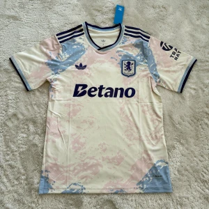 Aston Villa Adidas fotbollströja - Snygg Aston Villa fotbollströja från Adidas med Betano-tryck på bröstet. Tröjan är kortärmad och har ett unikt mönster i ljusblått och ljusrosa på en vit bas. Klassiska Adidas-ränder på axlarna och klubbmärke på bröstet. Tillverkad i lätt polyester med Aeroready-teknologi.