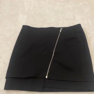 Svart kortkjol med dragkedja från H&M - Säljer en svart kortkjol från H&M i storlek 40. Kjolen har en snygg asymmetrisk design med en diagonal silverfärgad dragkedja framtill och dubbla lager nertill. Perfekt för dig som gillar stilrena och moderna plagg.