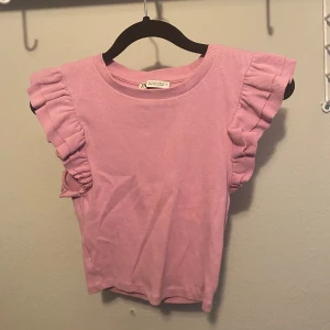 Rosa volangtopp från Zara - Supersöt rosa topp från Zara med volangdetaljer på axlarna. Toppen är ribbad och har en rund halsringning. Perfekt för dig som vill ha en feminin och trendig look. Materialet är mjukt och stretchigt, vilket gör den bekväm att bära.