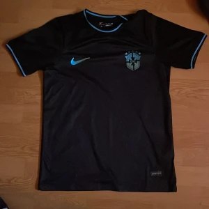 Svart Brasilien Nike fotbollströja - Snygg svart Brasilien fotbollströja från Nike med blå detaljer på krage och ärmslut. Tröjan har korta ärmar och är tillverkad i ett lätt, ventilerande material som passar perfekt för träning eller match. CBF-emblem och Nike-logga på bröstet.
