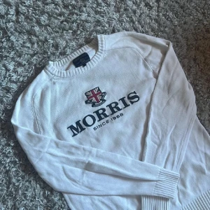 Vit stickad tröja från Morris - Vit stickad tröja från Morris med rund hals och broderad logga samt Union Jack-flagga på bröstet. Tröjan har långa ärmar och ribbade muddar vid hals, ärmslut och nederkant. Snygg och klassisk design med tydlig branding framtill.