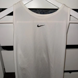 Vit Nike topp - Säljer en vit sportig linnetopp från Nike med Dri-Fit-teknologi. Toppen är ärmlös och har en klassisk rund halsringning samt svart Nike-logga framtill. Perfekt för träning och tillverkad i ett lätt och ventilerande syntetmaterial.