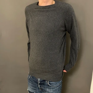 Mörkgrå stickad tröja i ull - En stilren mörkgrå stickad tröja med rund hals och långa ärmar. Tröjan är tillverkad i mjuk ull och har en klassisk, normal passform som funkar till jeans eller andra favoritplagg. Perfekt för dig som gillar enkel och clean stil.