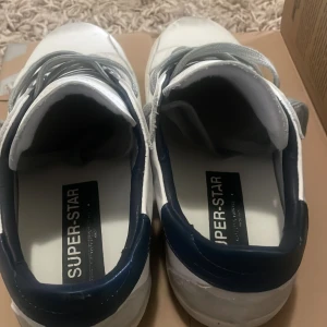 Vita sneakers med grå stjärna - Snygga vita sneakers från Super-Star med grå mockastjärna på sidan, grå snörning och mörkblå detalj vid hälen. Skorna har platt sula och är tillverkade i skinn med inslag av mocka. Perfekta för en trendig och avslappnad look.