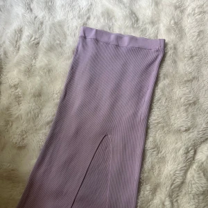 Ljuslila ribbad långkjol med slits - Säljer en ljuslila ribbad långkjol med hög midja och snygg slits framtill. Kjolen har en tight passform och är tillverkad i ett stretchigt material som sitter skönt på kroppen. Perfekt för dig som vill ha en stilren men ändå trendig look.