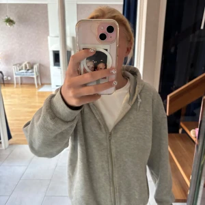 Grå hoodie med dragkedja från Gina Tricot - Mysig grå hoodie från Gina Tricot med dragkedja framtill och huva med snörning. Tröjan har långa ärmar, ribbade muddar och en relaxed passform. Perfekt för chill dagar eller när du vill vara extra bekväm.