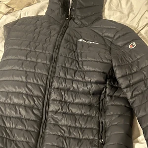 Svart pufferjacka från Champion S - Svart pufferjacka från Champion i storlek S. Jackan har huva, dragkedja framtill och två sidofickor med dragkedja. Quiltad design med diskret Champion-logga på bröstet och ärmen. Tillverkad i syntetmaterial med tech fill för extra värme.