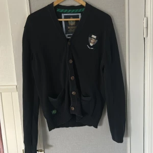 Marinblå kofta från Morris med emblem - Marinblå kofta från Morris med snyggt emblem på bröstet och knappar framtill. Klassisk v-ringad modell med två fickor och ribbade muddar. Perfekt för lager-på-lager och passar till många olika stilar.