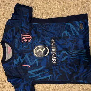 Atlético Madrid blå bortatröja Nike - Säljer en blå Atlético Madrid bortatröja från Nike med rosa detaljer och tryck. Tröjan har korta ärmar, klubbmärke på bröstet, sponsortryck och spelarnamn J. Álvarez med nummer 19 på ryggen. Materialet är lätt och andas, perfekt för fotbollsträning.