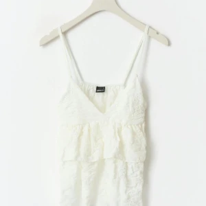 Vit volangtop från Gina Tricot - Supersöt vit linnetopp från Gina Tricot med tunna axelband och v-ringning. Toppen har dubbla volanger och ett lätt skrynklat tyg som ger en luftig känsla. Perfekt för varma dagar och enkel att styla med jeans eller kjol.