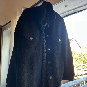 Svart teddyjacka med fickor - Svart teddyjacka med grov, fluffig yta och klassisk skjortkrage. Jackan har två stora bröstfickor med knappar och stängs med knappar framtill. Långärmad och loose fit, perfekt för dig som gillar en chill och mysig stil.