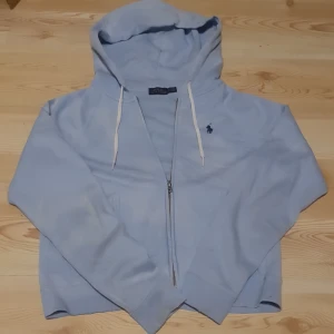Ljusblå hoodie från Polo Ralph Lauren XS/S - Säljer en snygg ljusblå hoodie från Polo Ralph Lauren med dragkedja och klassisk logga på bröstet. Perfekt för en avslappnad stil. Den har en justerbar huva med snören och långa ärmar och lite kort i midjan. Har några små hål dock. Ny pris över 1300kr
