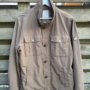 Field jacket från J.Lindeberg - Snygg oliv grön field jacka med ståkrage och fyra framfickor, två med lock och knappar. Jackan har knappstängning och justerbara detaljer i midjan. Perfekt för dig som gillar stilren och enkel design.