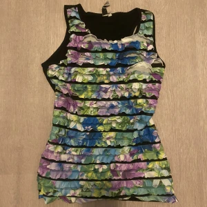 Blommig ärmlös linnetopp med volanger - Säljer en färgglad ärmlös linnetopp från Design Collection med blommigt mönster i blått, lila, grönt och vitt. Toppen har volanger i lager på framsidan och svart bakstycke. Perfekt för dig som gillar unika detaljer och somriga vibes.