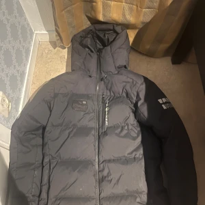 Svart jacka från Sail Racing - Svart pufferjacka från Sail Racing med huva och snygga loggor på ärmarna. Jackan har dragkedja framtill och quiltad design som ger en cool look. Perfekt för dig som vill ha en varm och stilren jacka med streetkänsla.