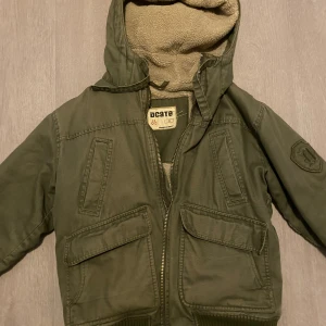 Olivgrön jacka med teddyfoder från Dcate - Olivgrön jacka från Dcate med mysigt teddyfoder och huva. Jackan har flera fickor framtill, dragkedja och en patch på ärmen. Perfekt för kyliga dagar och har en cool, avslappnad stil.