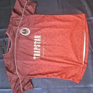 Trapstar vinröd fotbollströja S - Säljer en vinröd fotbollströja från Trapstar med beige tryck och nummer 22 på ryggen. Tröjan har korta ärmar, diskret mönster och vita detaljer längs axlarna. Tillverkad i lätt och ventilerande polyester, perfekt för dig som gillar streetwear och sportig stil.