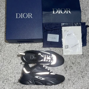Svarta Dior B22 sneakers med reflex - Christian Dior B22 sneakers i svart med reflekterande detaljer och meshpaneler. Snygg design med vita och silverfärgade inslag, grov sula och tydlig Dior-logga på sidan. Skorna har snörning och en modern, sportig vibe. Perfekta för dig som vill sticka ut med exklusiva sneakers.