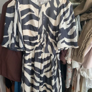 Zebramönstrad klänning med vida ärmar - Snygg klänning med zebramönster i svart och vitt. Modellen har vida, korta ärmar och markerad midja med rynkdetalj framtill. Lätt och luftig känsla, perfekt för dig som vill sticka ut med ett djurmönster.