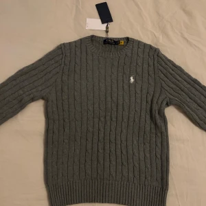 Grå kabelstickad tröja Polo Ralph Lauren - Snygg grå kabelstickad tröja från Polo Ralph Lauren i 100% bomull. Klassisk rund hals, ribbade muddar och vit broderad logga på bröstet. Perfekt för dig som gillar stilrena och tidlösa plagg med premiumkänsla.