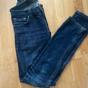 Mörkblå jeans från Nudie Jeans W26 L32 - Säljer ett par mörkblå jeans från Nudie Jeans med klassiska orange sömmar och snygga bakfickor med vågdetalj. Modellen har slim passform och raka ben. Tillverkade i slitstarkt bomullstyg, perfekta för en avslappnad stil.