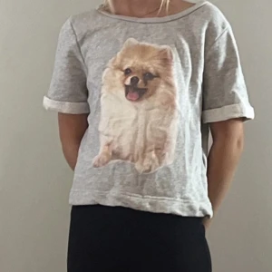 Grå t-shirt med hundtryck - Grå t-shirt i bomull med ett stort tryck av en söt pomeranian på framsidan. Modellen är kortärmad med uppvikta ärmslut och har en avslappnad passform. Perfekt för dig som älskar hundar och vill ha en chill look.